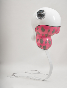 An organic oval shaped sculpture with a black cirlce where an eye would be and a loosely shaped oval in pink with grey patches of a textile adhered. A long white form supports the shape.
