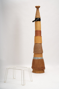 Multilayered totem-shaped sculpture made of multiple components joined together. A Small white plastic table like structure sits beside the work.