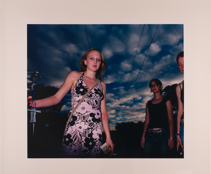 A young woman wears a halter neck styler dress decorated with black and white flowers with a bag strapped across her body. She looks down at the camera with two figures to her face right side. She is illuminated by a bright flash.