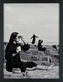 A black and white portrait orientated work that features three cloned female figures. Two are seated one is upright with her back to the camera. The figure in the foreground is holding a pair of binoculars and holding a half eaten sandwich in her right hand.