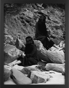 A black and white image of two female figures sit atop rocks on a sandy ground. Both figures have their faces obscured by their hair.