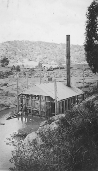 Tin shed with chimney on barge in water. Land and hills behind barge.