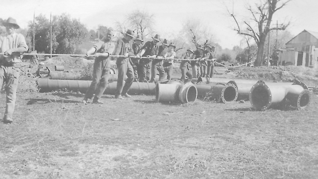 Eleven men pulling on cable, surrounded by sections of pipes.