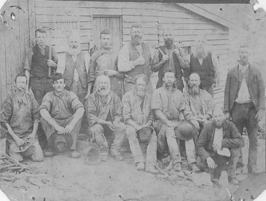 Fourteen men (seven sitting in front row, seven standing in back row), some holding tools, posing in front of slat timber building.