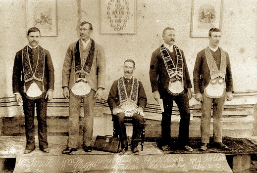 Five men on a raised platform - four standing, one sitting - wearing regalia of aprons and sashes.