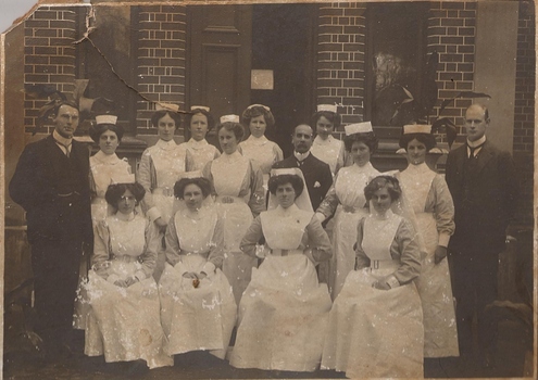 Twelve women in nurse uniforms (long sleeved with full length starched apron with bib, starched caps) and three men in suits posing at the entrance to a brick building