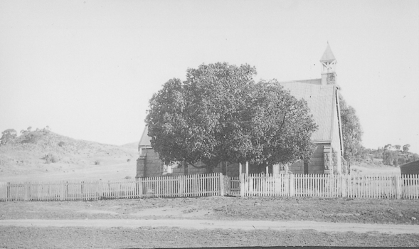 Large tree in front of brick building with tall roof and spire at front. Picket fence to the side of building.