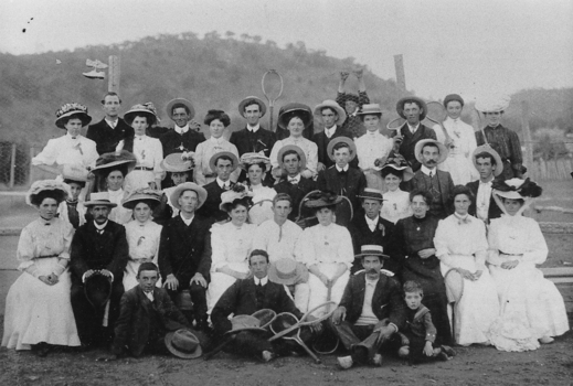 Group of men in suits and hats, women in long sleeved shirts, skirts, and bonnets, and one boy, with several tennis rackets.