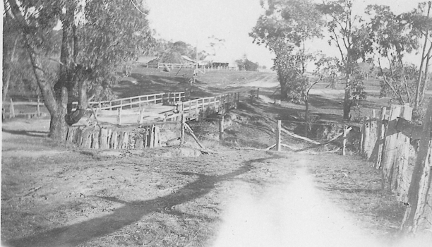 Bridge over a creek joining an unsealed road leading to a house on a rise overlooking the bridge.
