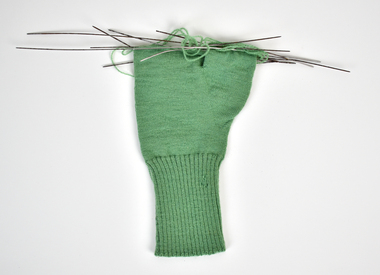 A green knitting glove in progress with knitting needles still attached