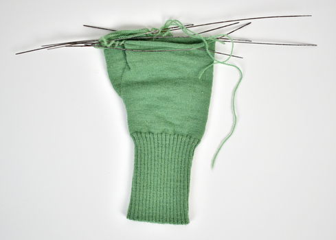 A green knitting glove in progress with knitting needles still attached