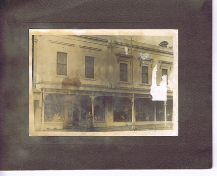View from street of shopfront of Miller Brothers China and Glass shop, in a row of shops. A woman stands in front of the doorway, under a verandah.
