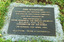 Rectangular metal plate inscribed with gold text and attached to concrete slab on the ground, surrounded by grass. The words on the plate are: This sculpture supersedes the original cenotaph located in the Catani Gardens and was unveiled by His Excellency Rear-Admiral Sir Brian Murray K.C.M.G., A.O., K.St.J., Governor of Victoria on Sunday, 21st April, 1985. Sculptor: Peter Schipperheyn 