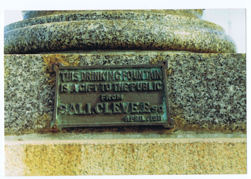 Metal plaque attached to base of granite pedestal. Raised letters on the plaque read 'This dining fountain is a gift to the public from Sali Cleve Esq April, 1911'