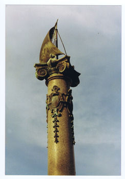 A bronze sculpture of a sailing ship sits on the scrolls on top of an ionic column. There are metal decorations on the column. 
