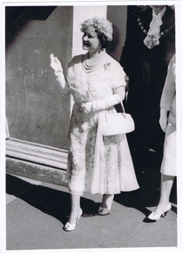 Smiling woman wearing a calf length frock, elbow length gloves, a hat and a pearl necklace carries a handbag over one arm and waves with the other.