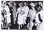 A crowd of women stand aside as they watch a woman holding a bouquet of flowers walk among them. All are wearing a calf length dress, a pair of gloves and a hat and are carrying a handbag over one arm.