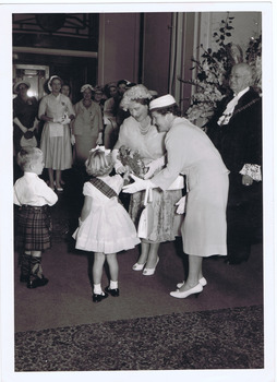 A young girl in frock hands a bouquet of flowers to a woman. Another woman and a young boy in a kilt watch on. A group of women and a man in ceremonial robes observe from a distance.