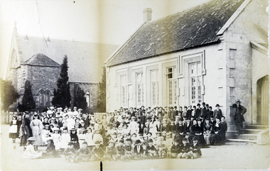 Dozens of children, and a few adults, sitting and standing in rows in front of a single storey brick building. In the background is a church.