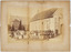 Dozens of children, and a few adults, sitting and standing in rows in front of a single storey brick building. In the background is a church.