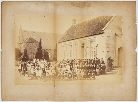 Dozens of children, and a few adults, sitting and standing in rows in front of a single storey brick building. In the background is a church.