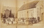 Dozens of children, and a few adults, sitting and standing in rows in front of a single storey brick building. In the background is a church.