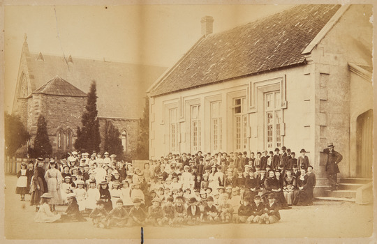 Dozens of children, and a few adults, sitting and standing in rows in front of a single storey brick building. In the background is a church.