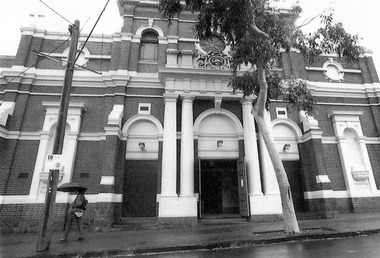 Dark brick building with contrasting white ornamentation. At ground level are three arched doorways. The main entrance is the middle doorway, which is framed by a portico supported on each side by two columns. Above the main entrance is a round stained glass window. On either side of the round window is an arched window.