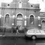 Brick building with contrasting white ornamentation. The main entrance is an arched doorway with a wooden door. Above the door is a sign and a semi-circular stained glass window. The sign says Sacred Heart Mission. On either side of the door is an arched window. On the right of the door is another sign, which says Sacred Heart Mission Op shop. Further to the right, and partially obscured, is another arched doorway and wooden door. Above the main entrance are three round windows. There is a cross on top of the roof. A man is standing in front of the main entrance with both hands raised. A car is parked in front of the building.
