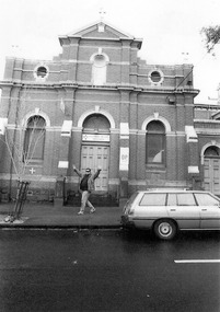 Brick building with contrasting white ornamentation. The main entrance is an arched doorway with a wooden door. Above the door is a sign and a semi-circular stained glass window. The sign says Sacred Heart Mission. On either side of the door is an arched window. On the right of the door is another sign, which says Sacred Heart Mission Op shop. Further to the right, and partially obscured, is another arched doorway and wooden door. Above the main entrance are three round windows. There is a cross on top of the roof. A man is standing in front of the main entrance with both hands raised. A car is parked in front of the building.