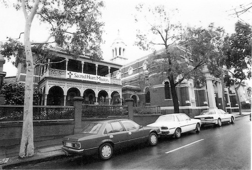 Two storey brick building with wrought iron verandah supported by columns and decorated with lacework. In front of the building is a brick and wood fence. Three cars are parked on the street. To the right of the building is a brick church with a white steeple.