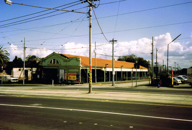 Long single storey building with red roof that extends into a verandah seen at an angle from across a street. The verandah covers a waiting area for trams. Tram tracks run beside the waiting area and along the street.