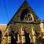 Stone church building with a high roof and walls supported by stone buttresses. There is a pointed arch door and a pointed arch stained glass window on each side of the door. Above them is a large triangular shaped window. The stained glass of the window is held by stone tracery that forms a triangle. Within the triangle the tracery forms a large circle that touches each of the triangle's sides. Within the large circle are four small circles, and within each small circle are four smaller circles. 