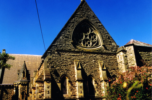 Stone church building with a high roof and walls supported by stone buttresses. There is a pointed arch door and a pointed arch stained glass window on each side of the door. Above them is a large triangular shaped window. The stained glass of the window is held by stone tracery that forms a triangle. Within the triangle the tracery forms a large circle that touches each of the triangle's sides. Within the large circle are four small circles, and within each small circle are four smaller circles. 