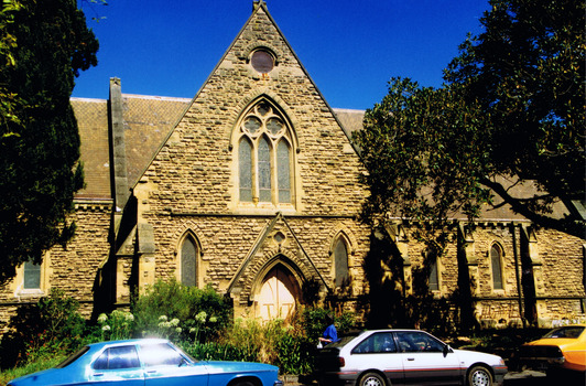 Stone church building with a high roof and walls supported by stone buttresses. At ground level are five pointed arch stained glass windows and a pointed arch wooden door. Above the doorway is a large pointed arch stain glass window. The stained glass is held by stone tracery that forms three pointed arches and three circles. 