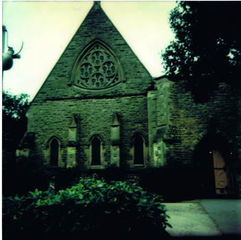 Stone church building with a high roof and walls supported by stone buttresses. There are three pointed arch doors and on their left a pointed arch wooden door. Above them is a large triangular shaped window. The stained glass of the window is held by stone tracery that forms a triangle. Within the triangle the tracery forms a large circle that touches each of the triangle's sides. Within the large circle are four small circles, and within each small circle are four smaller circles.
