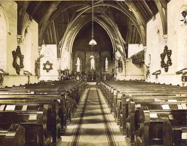 Rows of wooden pews on either side of a carpeted aisle that leads to an altar under a pointed arch. Three large wreaths of flowers in the shape of stars decorate the walls. Other flower arrangements are on the window sills and at the entrance to the altar.