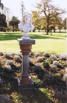 White marble bust of a woman on top of a grey and brown column and grey plinth. The statue is standing in a garden bed. Green lawn and trees are in the background. 