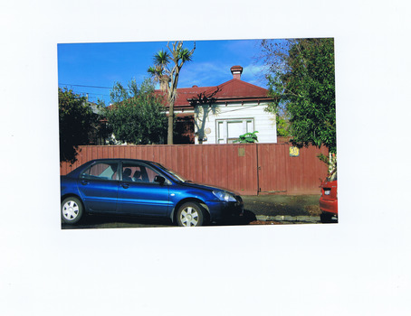 A blue car parked on the road in front of a brown wooden fence. Attached to the fence is a plaque and the number 3. Behind the fence are two trees, a shrub, and a white weatherboard house with a red corrugated iron roof.