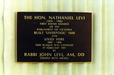 Rectangular metal plate inscribed with gold text and attached to a cream painted corrugated fence.