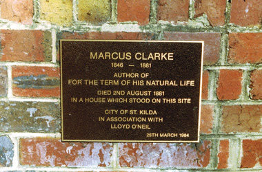 Rectangular metal plate inscribed with gold text and attached to a brick wall. 