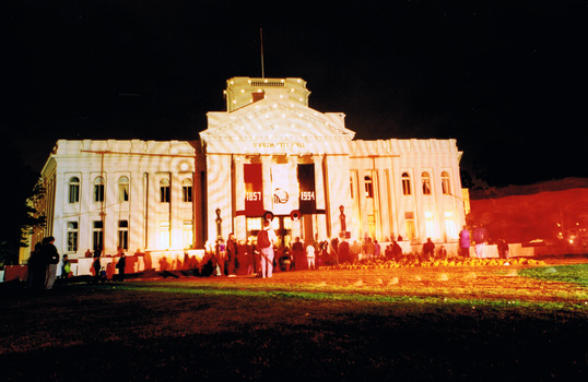 Floodlit two storey heritage building with a grand portico displaying the words St Kilda Town Hall. Three banners hang from the portico. The banner on the left reads 1857. The banner on the right reads 1994. The banner in the middle displays the City of St Kilda logo. 