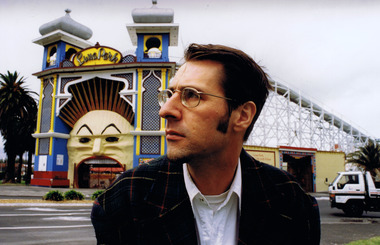Profile of head and shoulders of man wearing glasses, white shirt and dark checked jacket. The Luna Park entrance and scenic railway are in the distance behind him. 