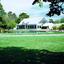 Pond surrounded by lawns, some trees and paths. In the distance is a building with windows along all sides and a high pitched glass roof. Two people stand near the pond. 