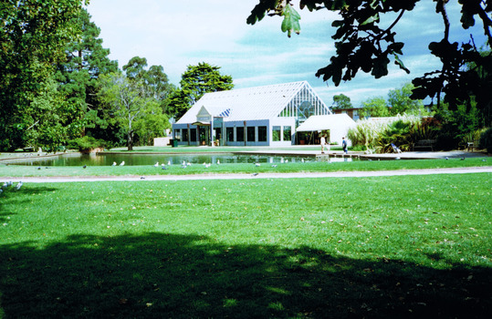Pond surrounded by lawns, some trees and paths. In the distance is a building with windows along all sides and a high pitched glass roof. Two people stand near the pond. 