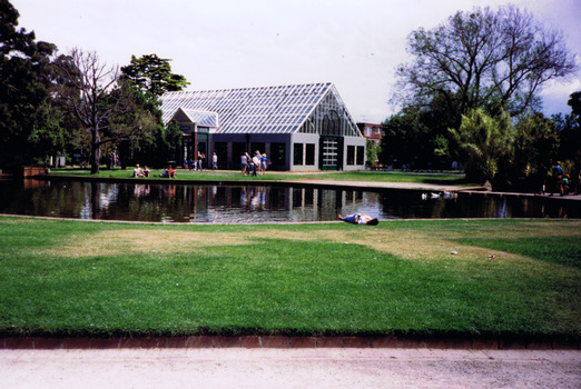 Pond with a brick border, surrounded by lawns and some trees. Some ducks swim in the water. In the distance is a building with windows along all sides and a high pitched glass roof. Groups of people sit and stand near the pond. One person lies down on the grass.