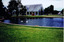 Pond with a brick border, surrounded by lawns and some trees. Some ducks swim in the water. In the distance is a building with windows along all sides and a high pitched glass roof. Groups of people stand nearby. 