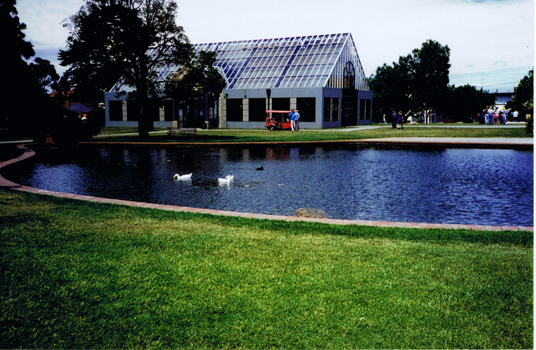 Pond with a brick border, surrounded by lawns and some trees. Some ducks swim in the water. In the distance is a building with windows along all sides and a high pitched glass roof. Groups of people stand nearby. 