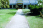 A grey gravel path leads to the entrance door of a building which has windows along the side and a high pitched roof. On either side of the path is grass. There is a portico over the entrance door and it displays the words St Kilda Conservatory.