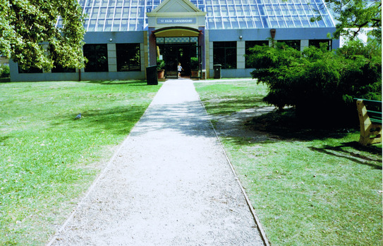 A grey gravel path leads to the entrance door of a building which has windows along the side and a high pitched roof. On either side of the path is grass. There is a portico over the entrance door and it displays the words St Kilda Conservatory.